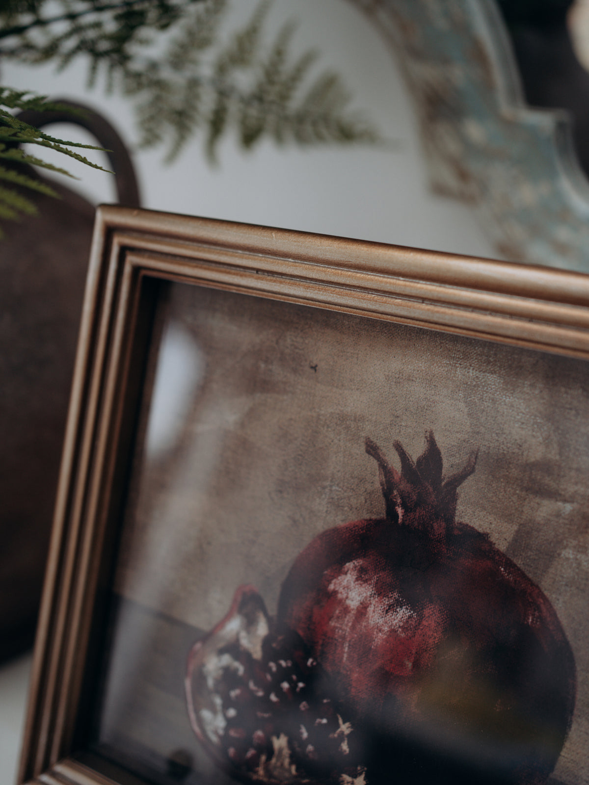 A framed still life artwork depicting a pomegranate, shown at an angle with a distressed gold frame and a softly blurred background.
