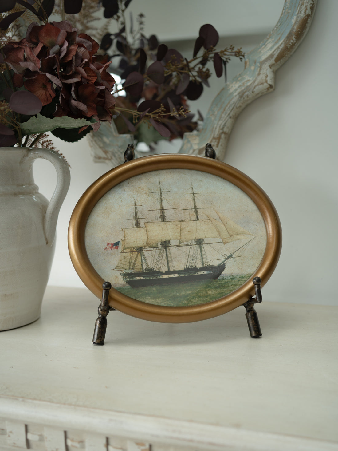 An oval framed decorative art featuring a painted sailing ship, displayed on a white mantel with a ceramic vase and dried flowers in the background.