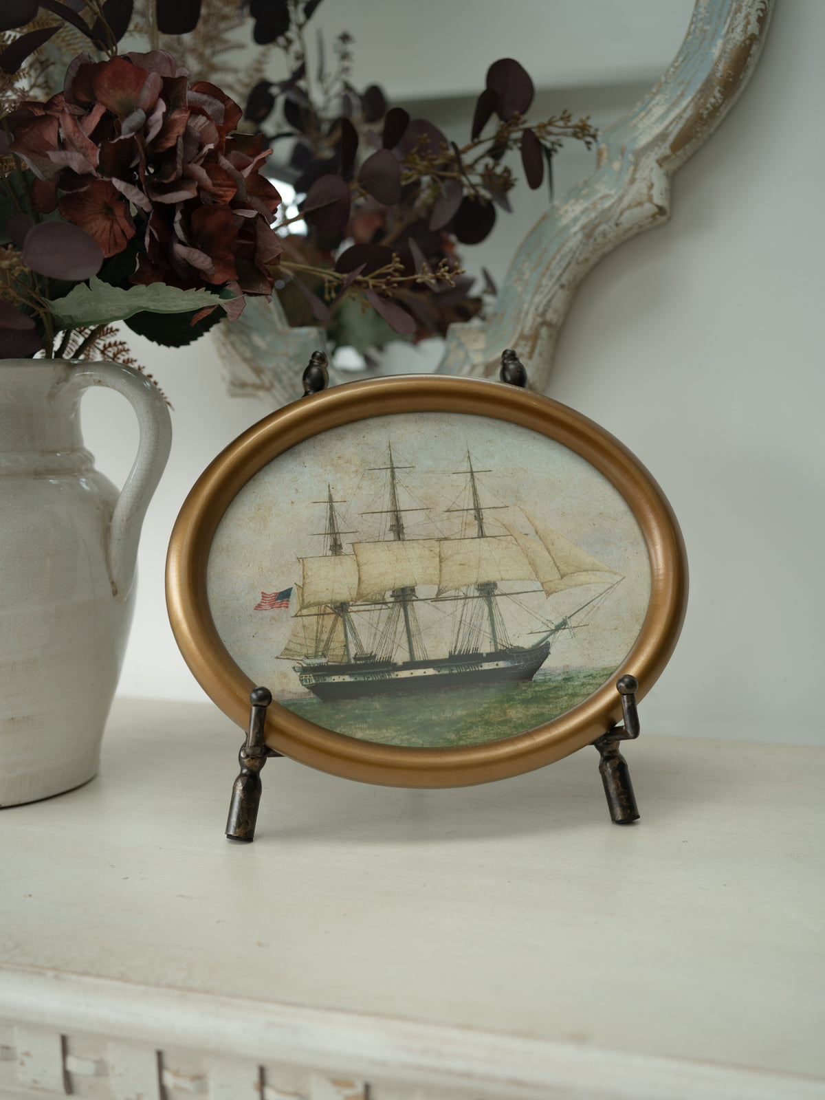 An oval framed decorative art featuring a painted sailing ship, displayed on a white mantel with a ceramic vase and dried flowers in the background.