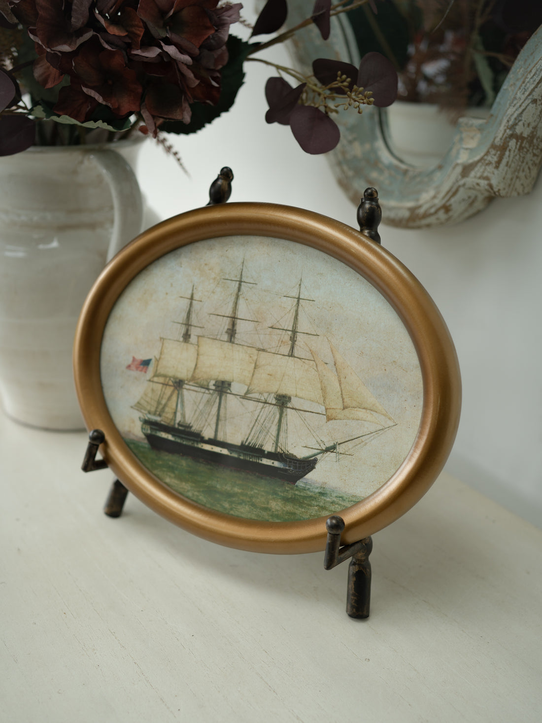 An oval framed decorative art featuring a vintage sailing ship illustration, displayed on a tabletop with a small dark metal stand. The frame is a warm metallic gold, and the background includes a white vase with dark dried flowers and a distressed mirror.