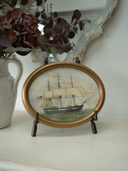 An oval framed miniature painting of a historic sailing ship on the water, displayed on a small dark metal stand on a white shelf next to a ceramic vase with dried flowers.