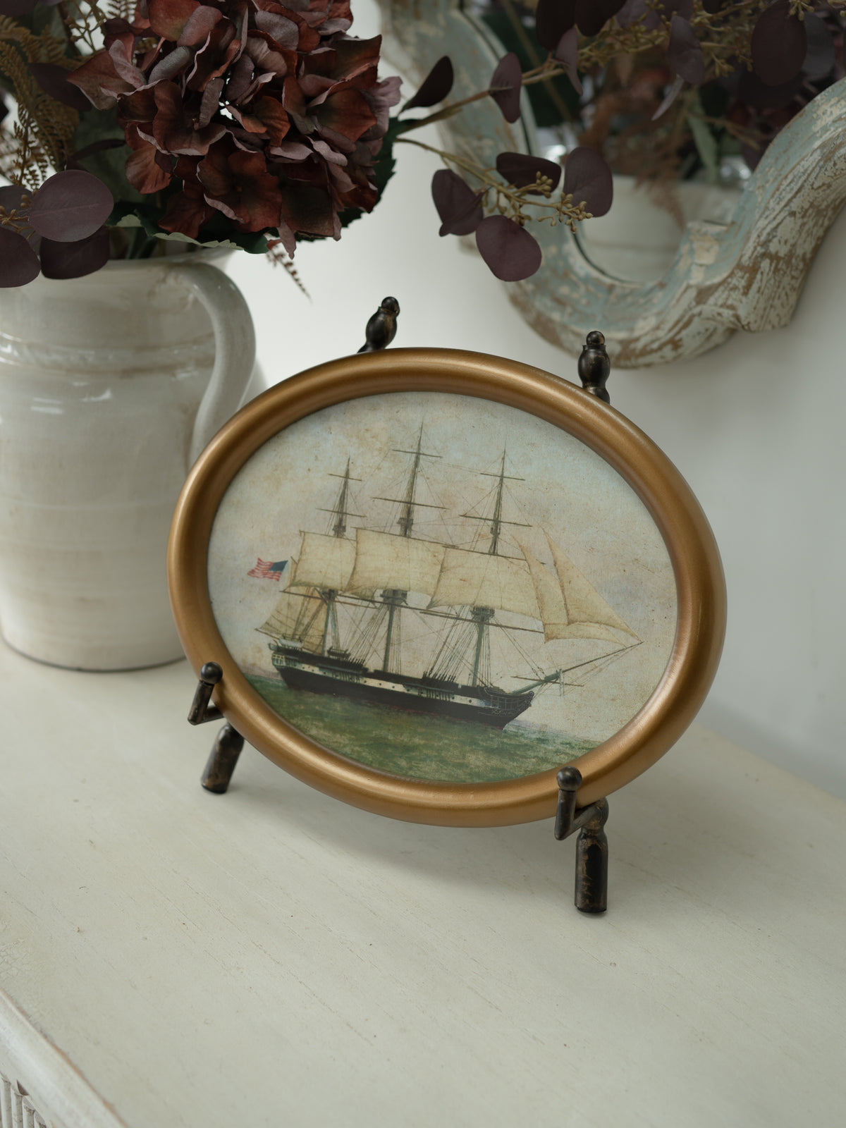 An oval framed art print featuring a vintage sailing ship, displayed on a small black stand on a white tabletop with decorative dried flowers and a mirror in the background.