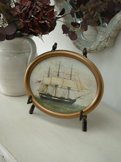 An oval framed art print featuring a vintage sailing ship, displayed on a small black stand on a white tabletop with decorative dried flowers and a mirror in the background.