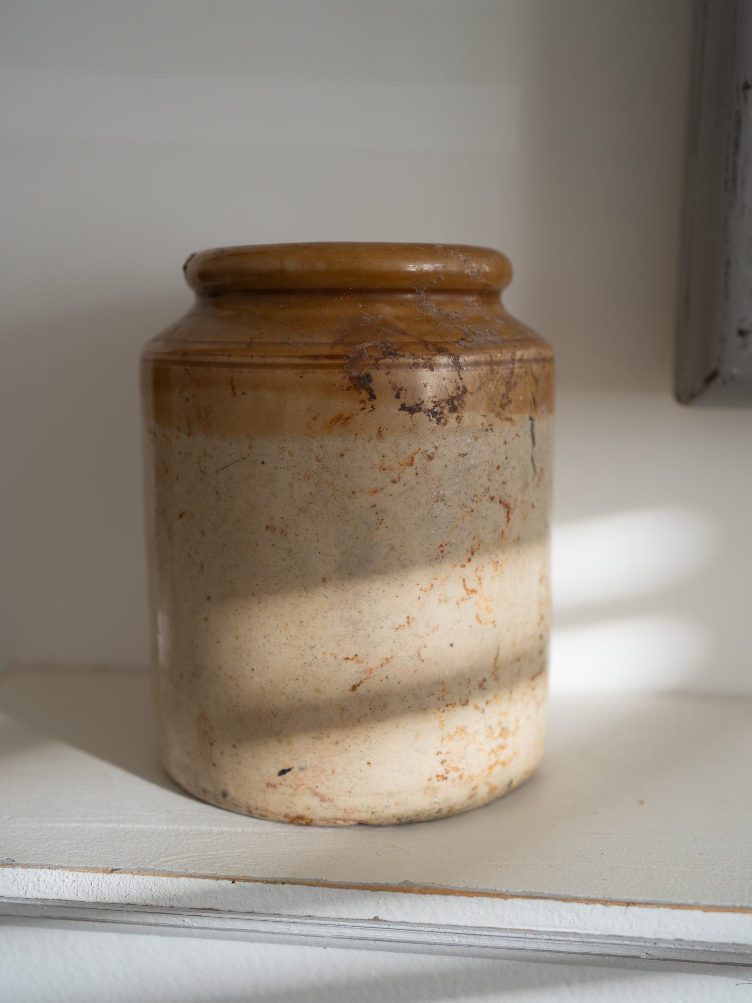 A rustic ceramic storage jar with a brown glazed top and a speckled beige lower body, sitting on a white shelf in soft natural light.