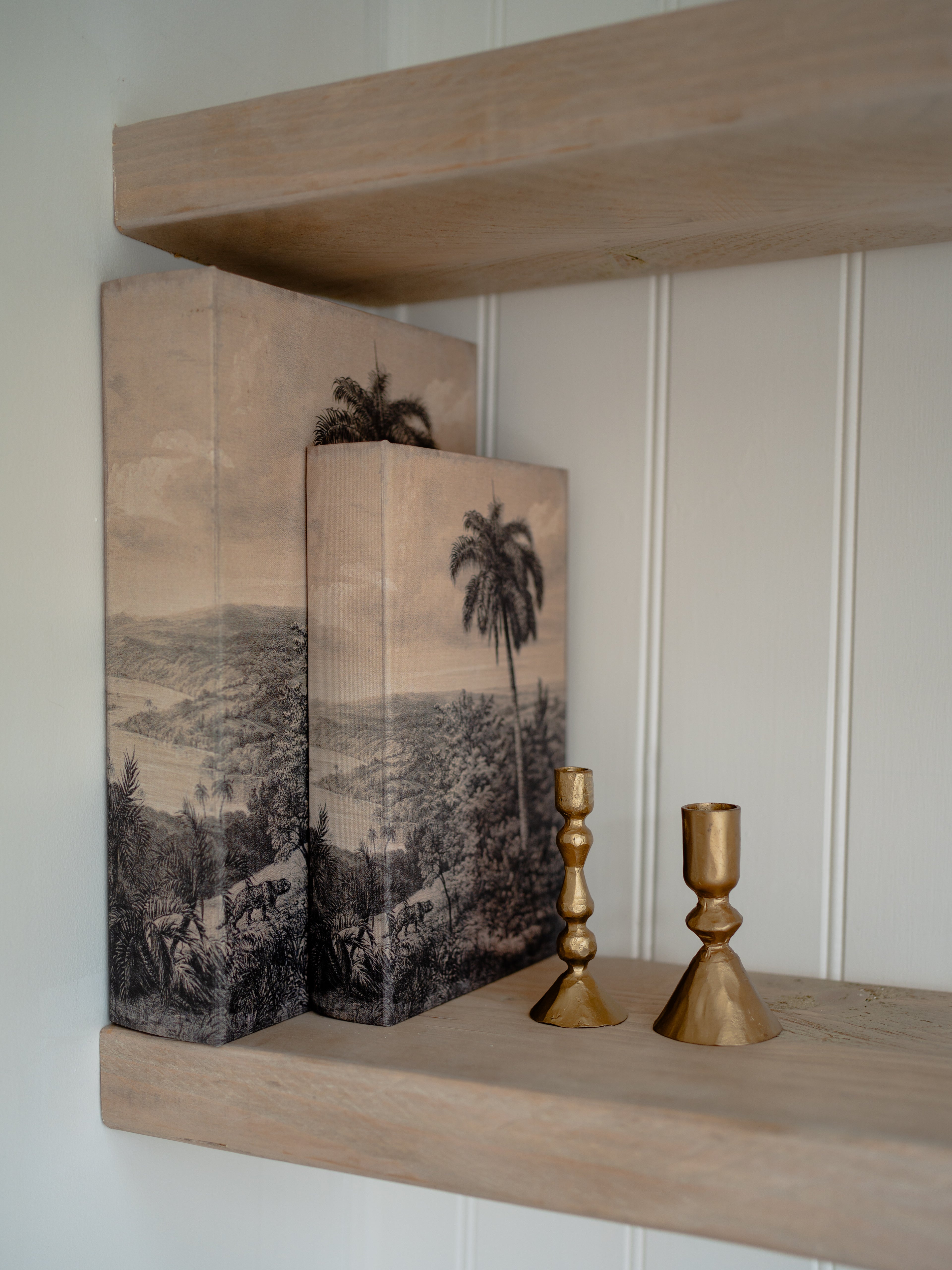Decorative storage book boxes with landscape illustrations on a wooden shelf with gold candle holders.