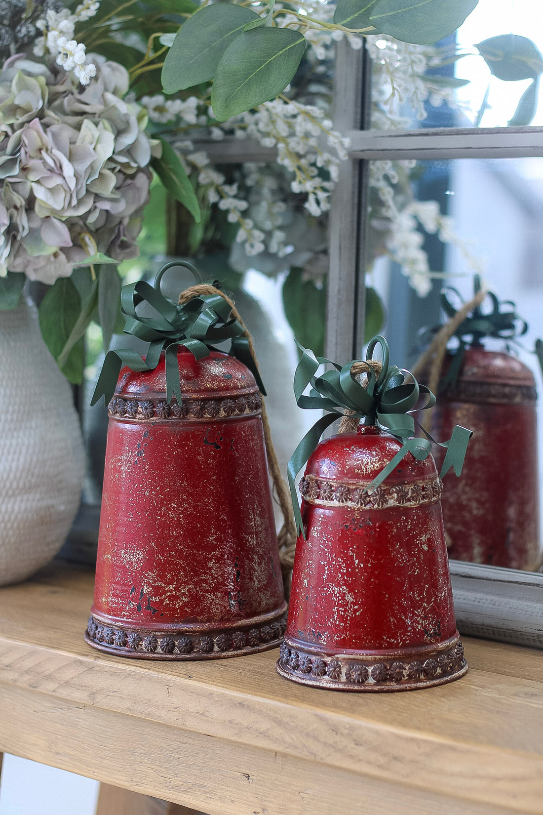 Two red decorative bells with green ribbons on a wooden surface with flowers in the background.