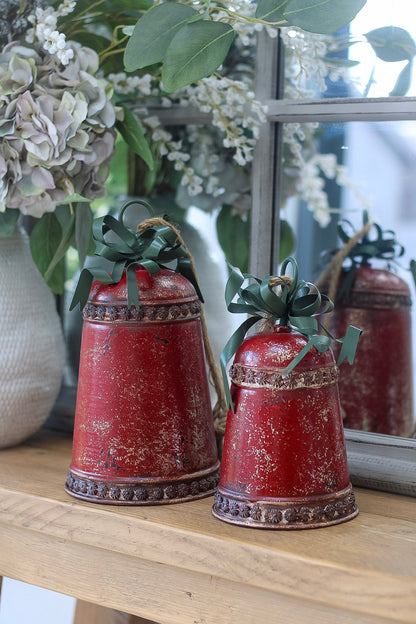 Two red decorative bells with green ribbons on a wooden surface with flowers in the background.
