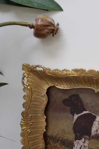 Decorative gold frame with a painting of a dog on a white surface with a dried flower.