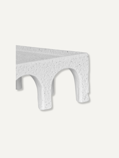 A close-up of a white textured stone or concrete architectural detail with a rough, speckled finish and a curved arch-like recess, shown against a solid off-white background.