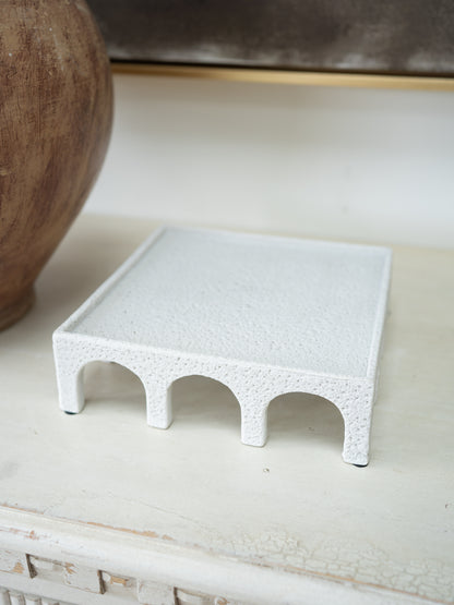 A small white textured tray with a raised border and three arch openings underneath, styled on a distressed white surface next to a wooden vase.