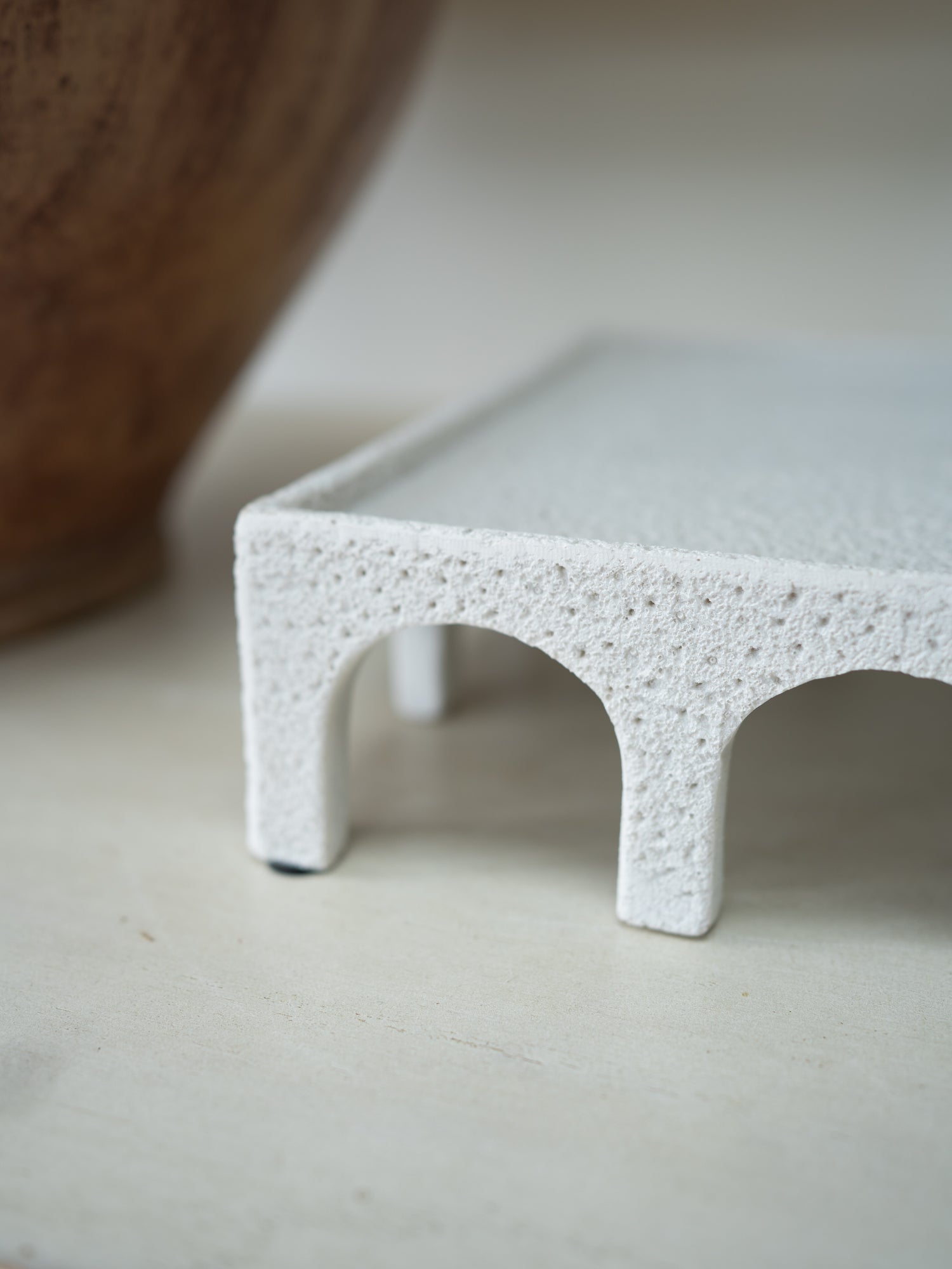 A close-up of a small white textured ceramic stand with two arches and a flat rectangular top, photographed on a light neutral surface.