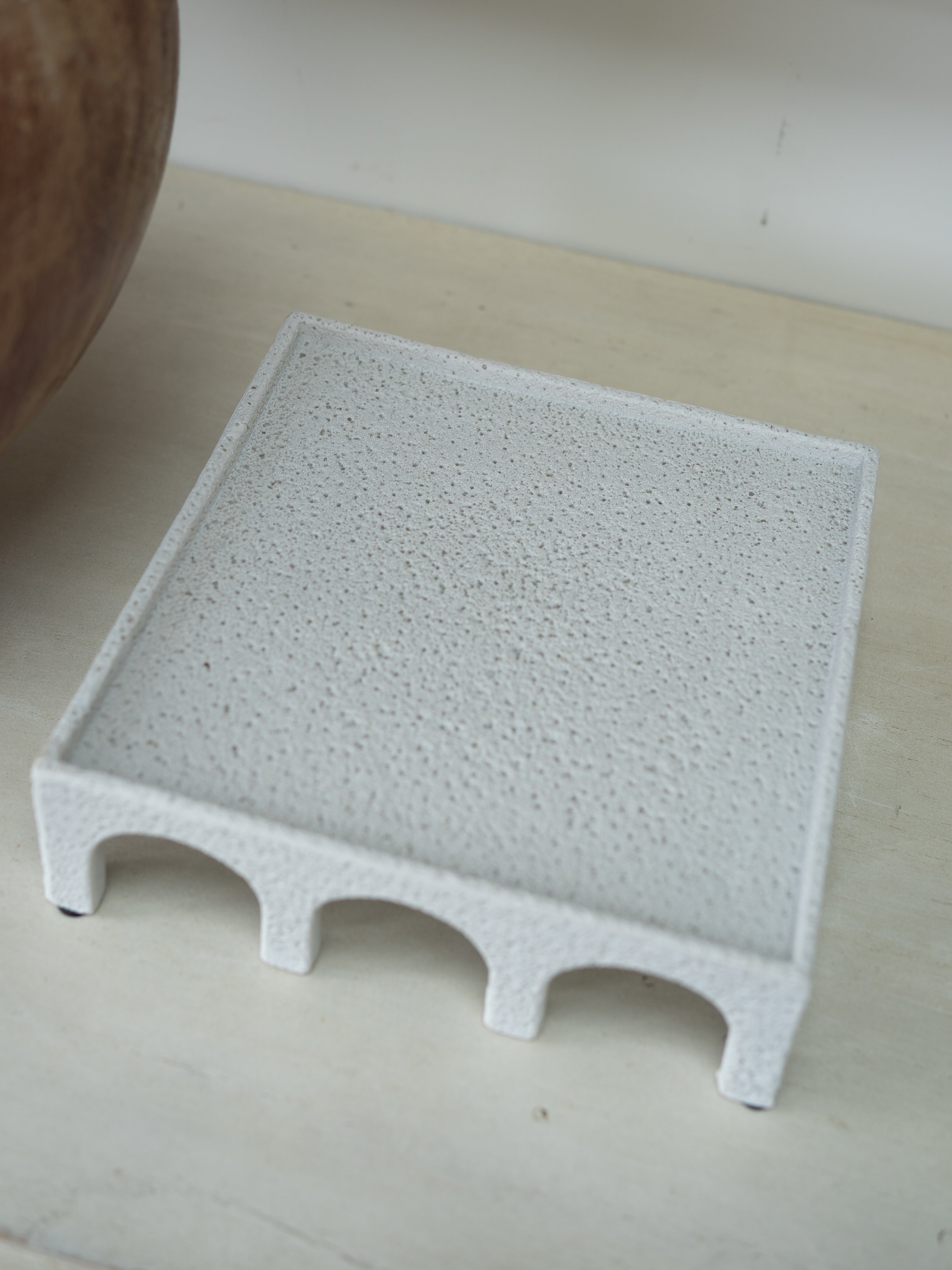 A square white textured tray with a raised rim and three arch-shaped feet along the bottom, photographed on a light beige surface.