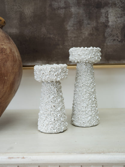 Two white textured decorative candle holders with a bubbly, coral-like surface finish, displayed on a light surface next to a wooden vase against a neutral background.