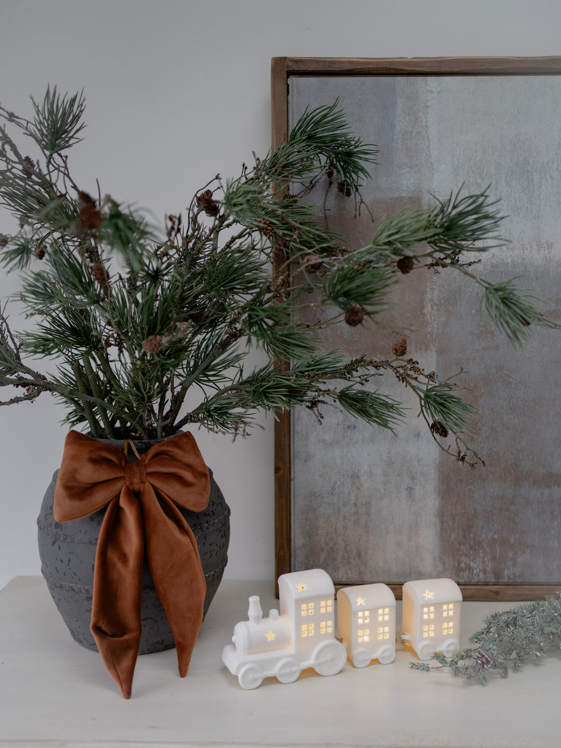 Decorative setup with a vase of greenery, a brown bow, and small white houses on a white surface.