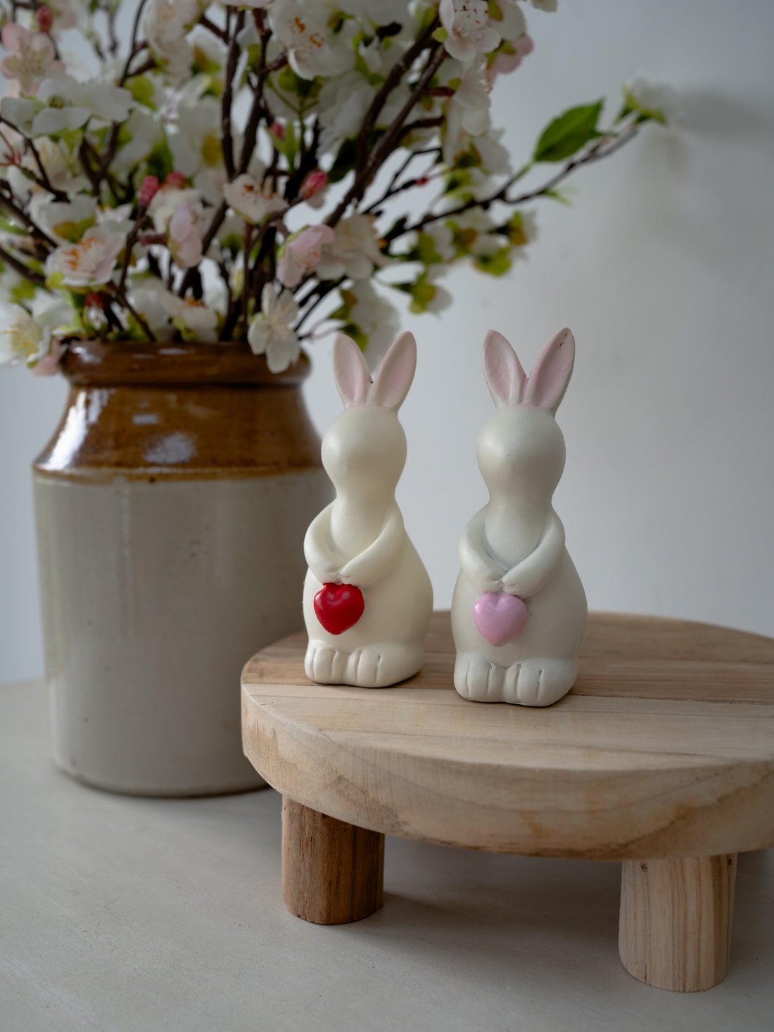 Two ceramic bunny figurines holding hearts on a wooden stand with a vase of flowers in the background.