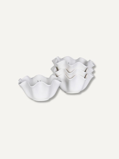 Four white ceramic ruffled bowls, with three stacked and one standing, on a light beige background.