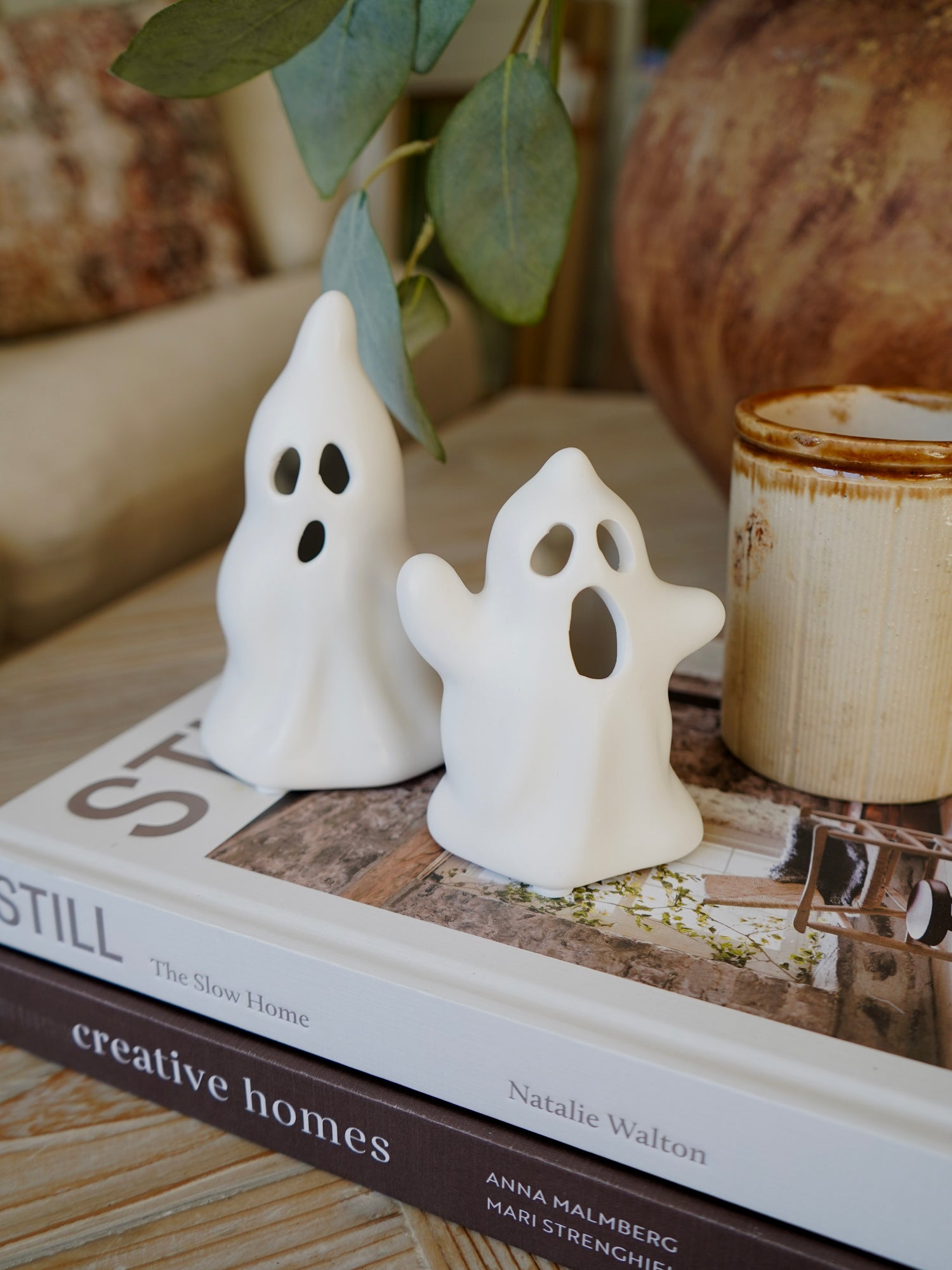 Two small white ghost figurines on a book with a candle and plant in the background
