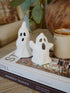 Two small white ghost figurines on a book with a candle and plant in the background