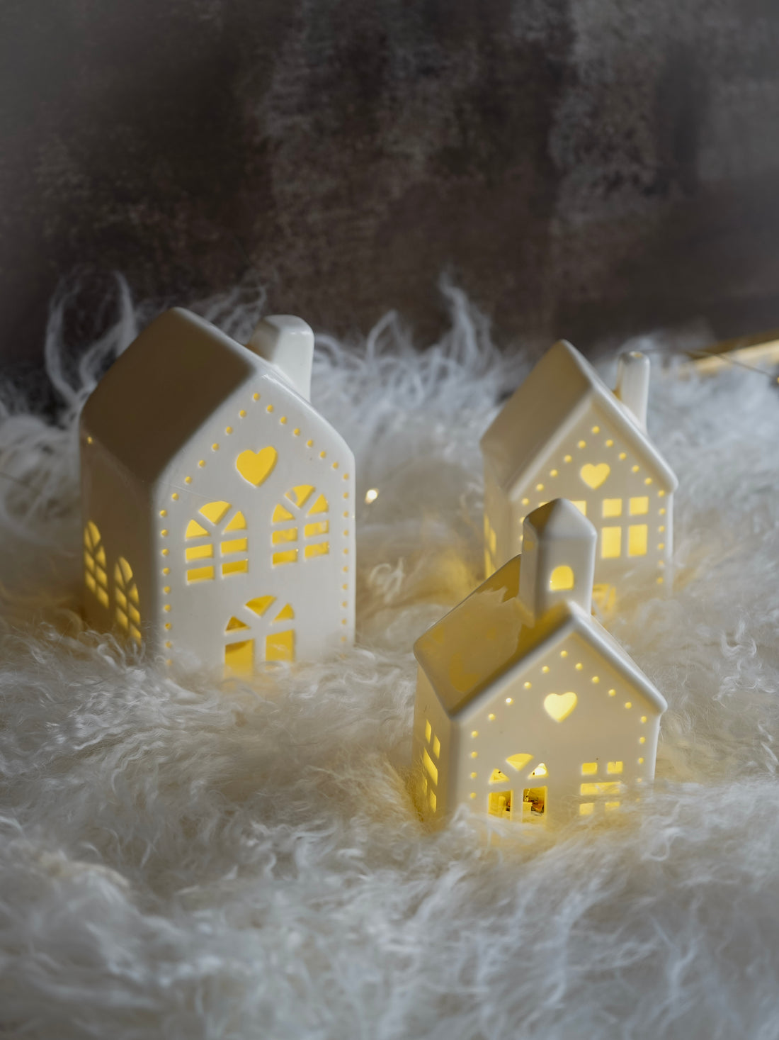 Three illuminated white house-shaped lanterns on a soft surface with a blurred natural background