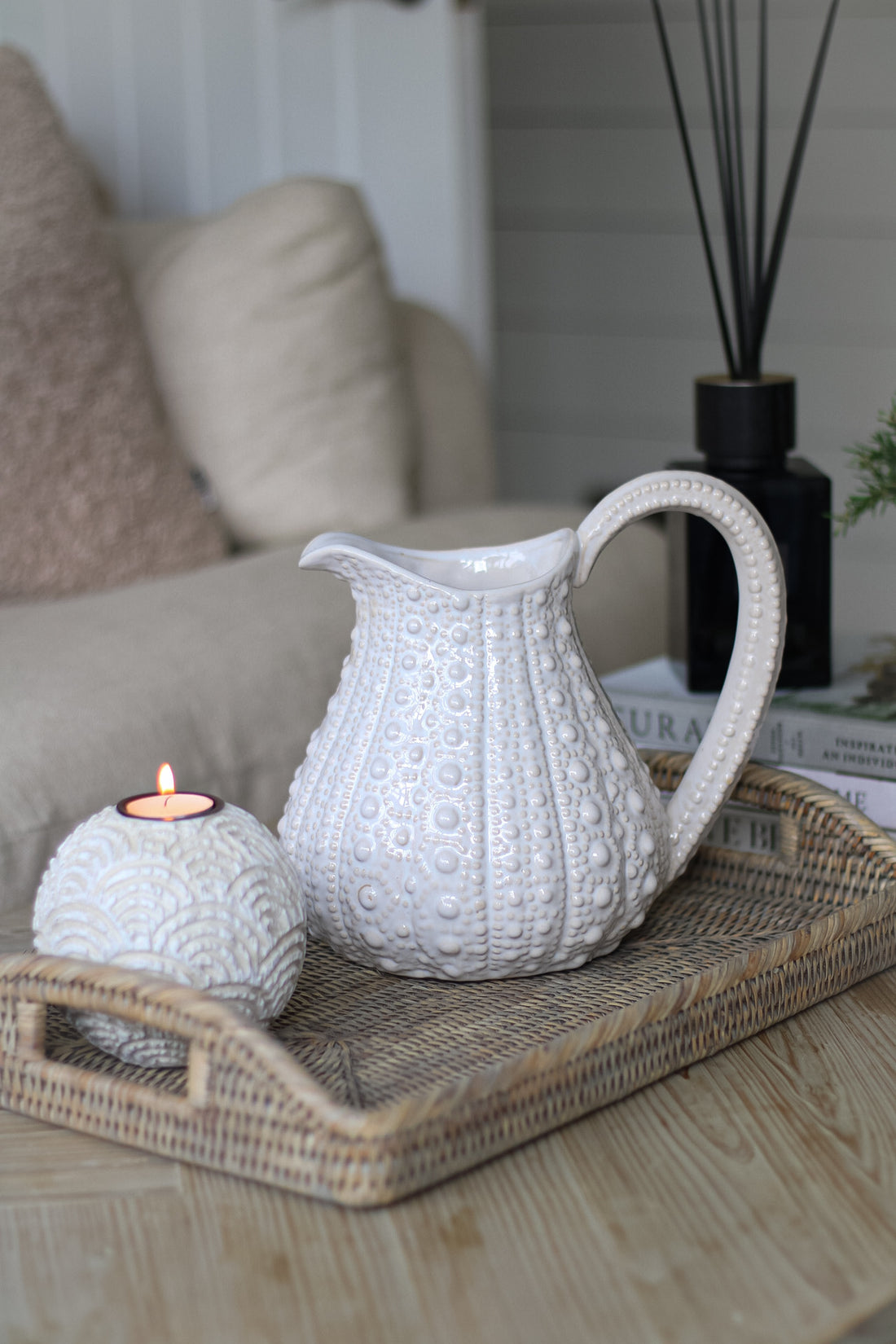 A white porcelain sea urchin jug with a textured design, placed on a wicker tray on a living room table, accompanied by a small round object that could be a candle holder.
