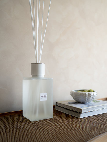 A frosted glass reed diffuser bottle with white reeds and a small front label reading SENCES, styled on a woven wooden beside stacked books and a textured bowl against a warm beige wall.