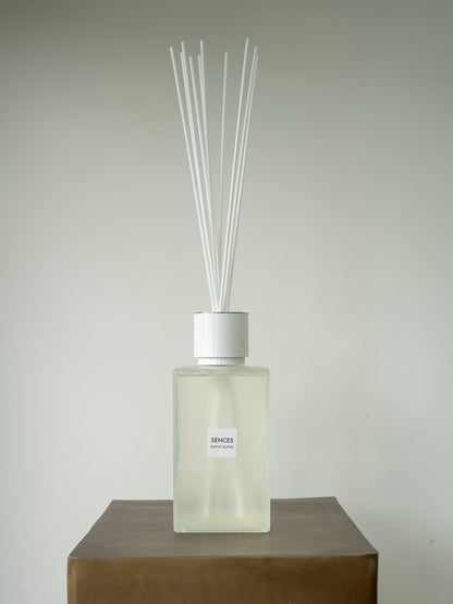 A sences alang alang reed diffuser bottle with a frosted glass body, white cap, and multiple white reeds, displayed on a brown block against a light gray background.