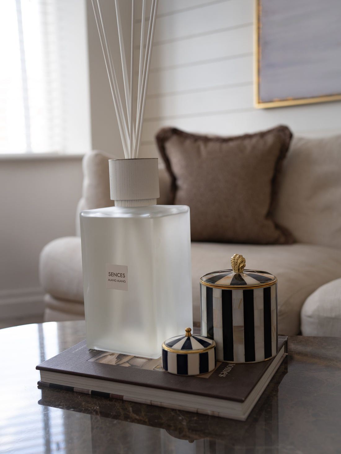 A frosted glass reed diffuser bottle with white reeds stands on a tray coffee table two black-and-white striped decorative boxes with gold trim in a softly lit living room setting.