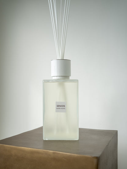 A reed diffuser bottle with a frosted glass body, white cap, and white reeds, labeled &
