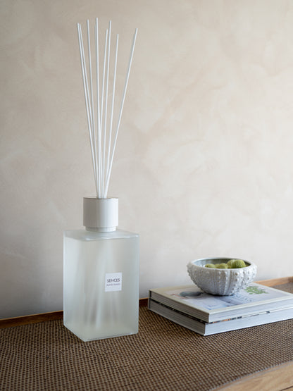 A frosted glass SENCES ALANG ALANG reed diffuser bottle with white reeds stands on a woven mat next to a stack of books and a small textured bowl, against a warm beige wall.