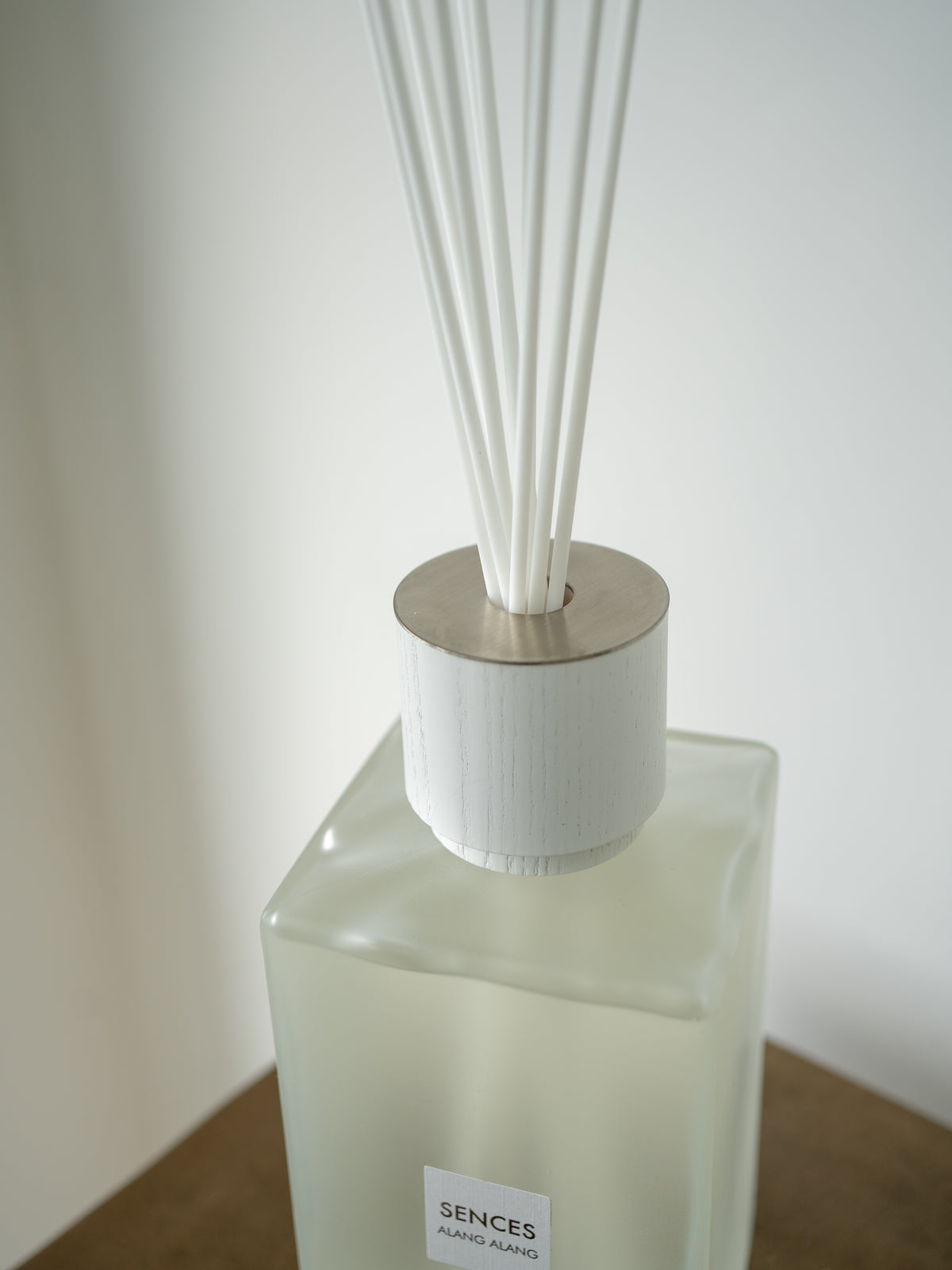A close-up of a SENCES reed diffuser bottle with a frosted glass body, white reeds, and a brushed silver metal top, resting on a wooden surface against a neutral background.