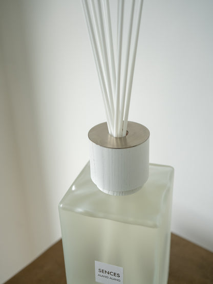 A close-up of a SENCES reed diffuser bottle with a frosted glass body, white reeds, and a brushed silver metal top, resting on a wooden surface against a neutral background.