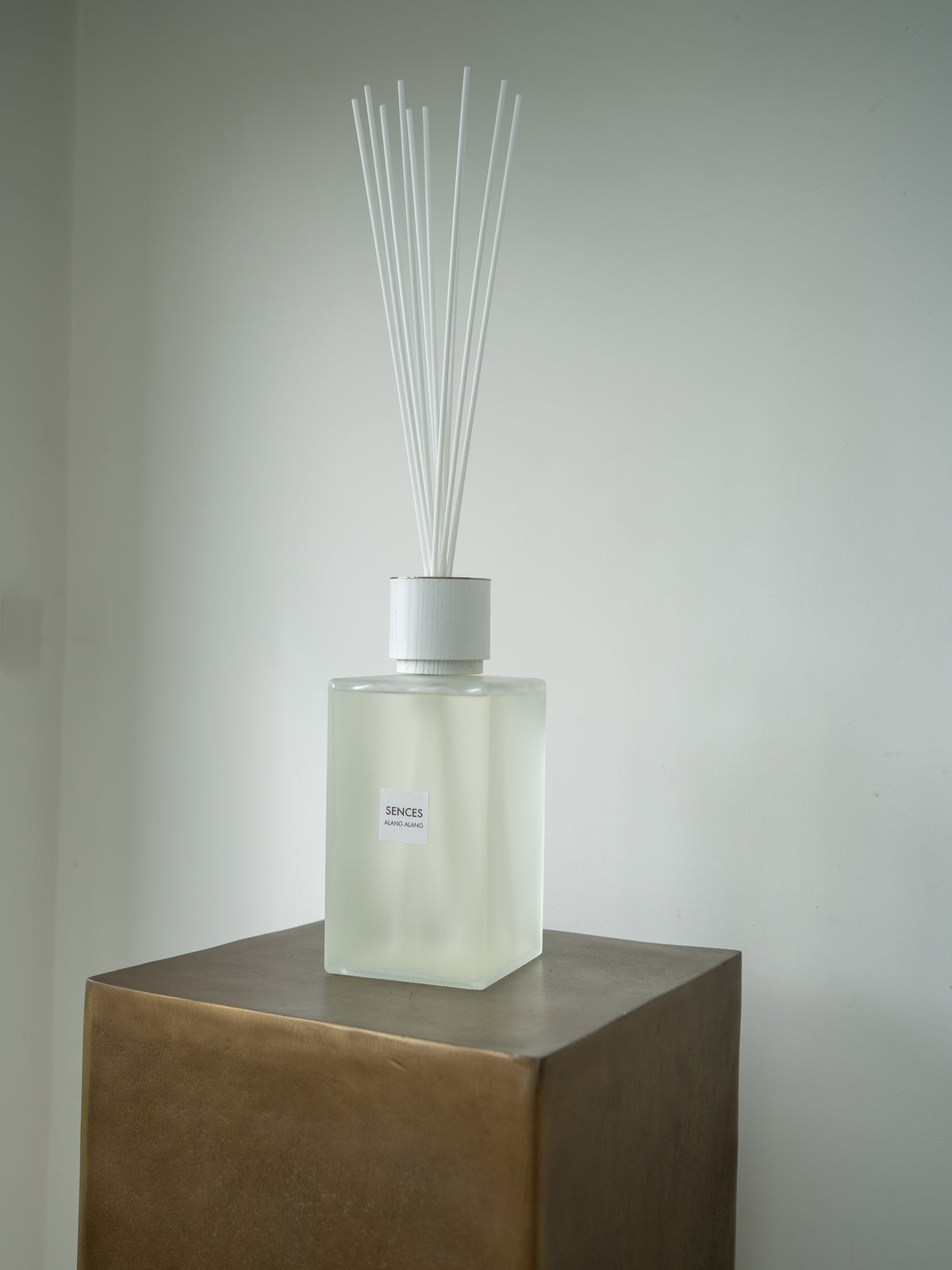 A sences alang alang reed diffuser bottle with white reeds, placed on a metallic bronze pedestal against a plain light gray wall.