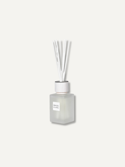 A frosted glass reed diffuser bottle with a white cap and clear reeds, centered on a light off-white background. The label reads &