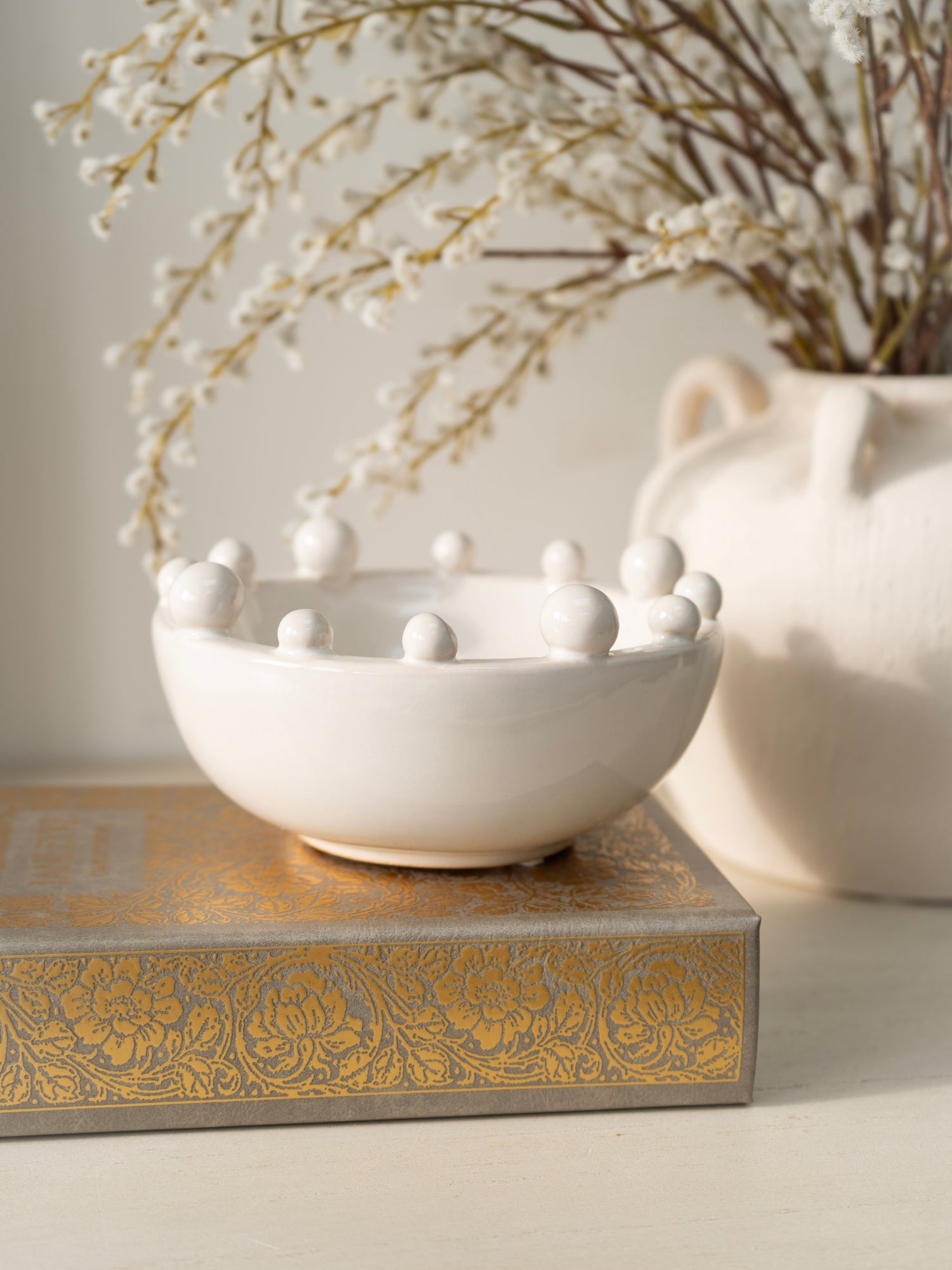 White ceramic bowl with spherical bobble decorations on a decorative box, with a vase of branches in the background.