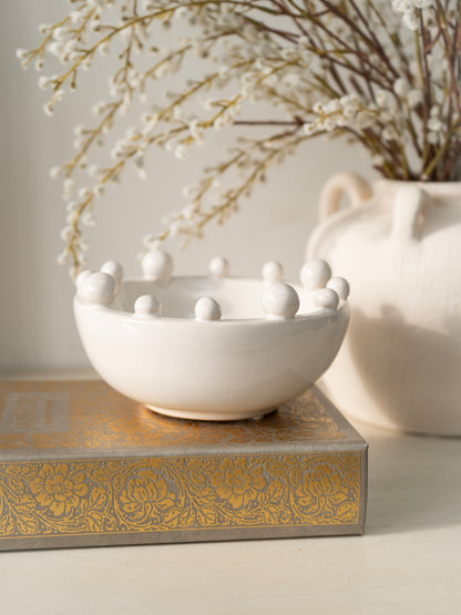 White ceramic bowl with spherical bobble decorations on a decorative box, with a vase of branches in the background.