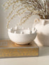 White ceramic bowl with spherical bobble decorations on a decorative box, with a vase of branches in the background.