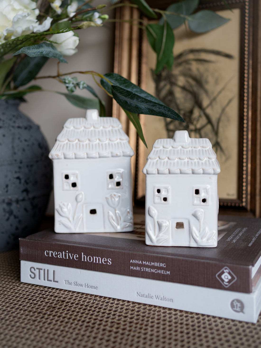 Two white ceramic house-shaped candle holders on a stack of books with decorative elements in the background.