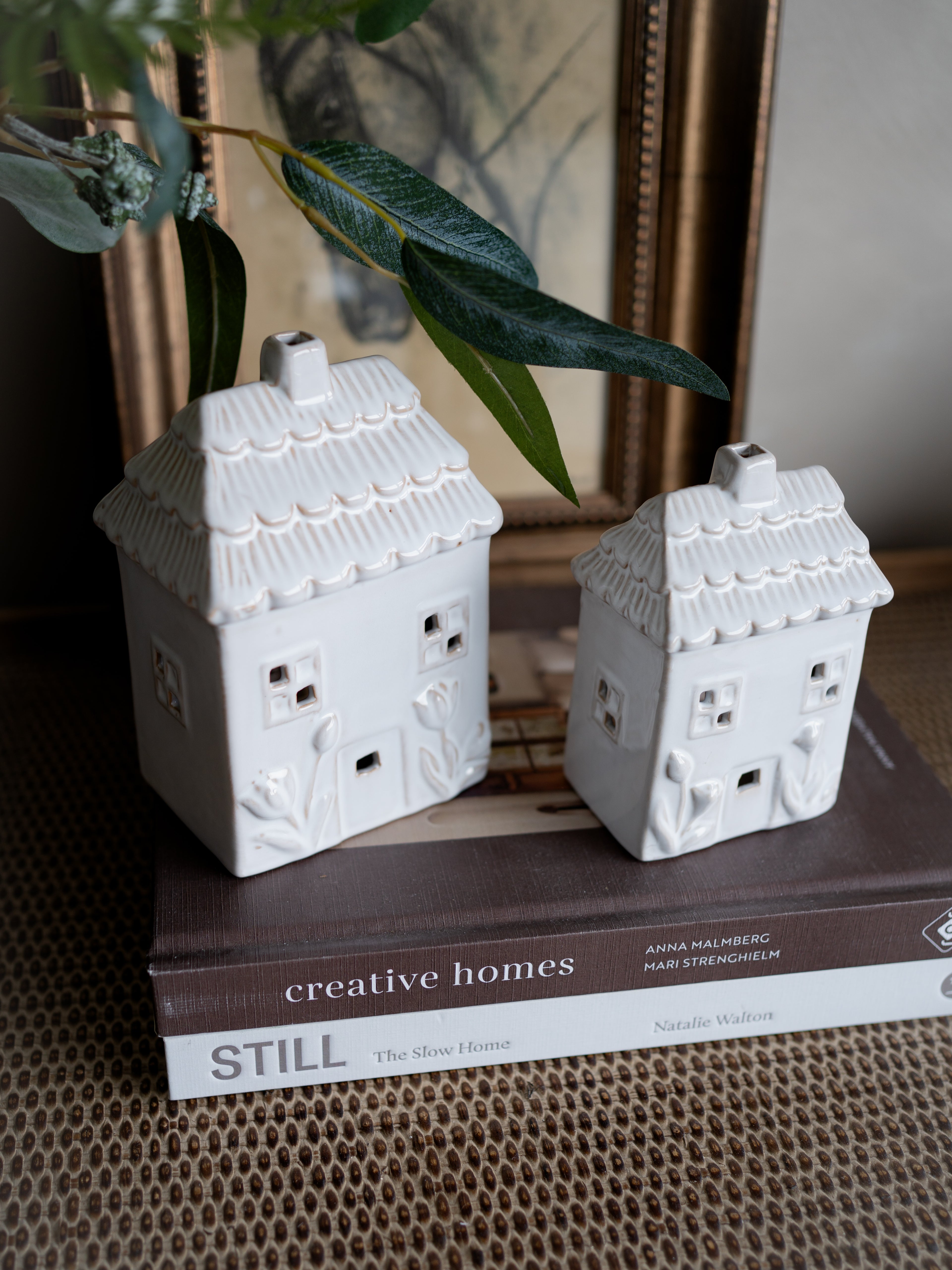 Two white ceramic house-shaped objects on a book with a plant in the background.