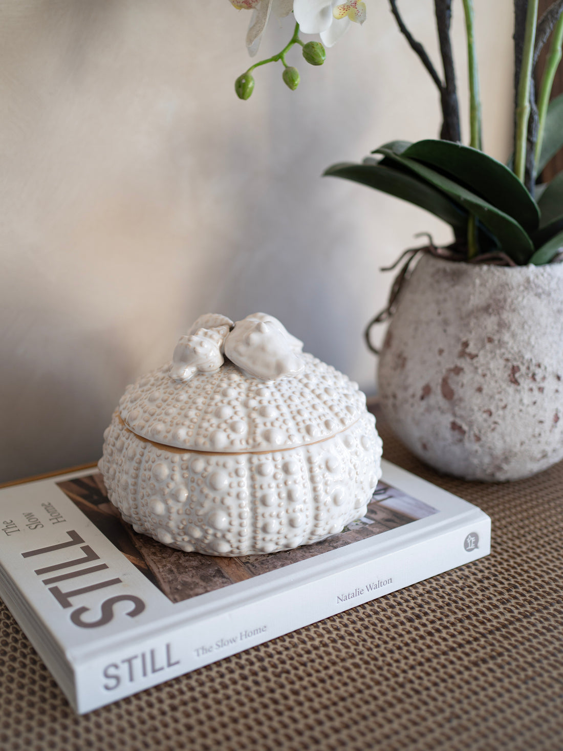 Decorative white textured urchin jar on a book with a vase of flowers in the background