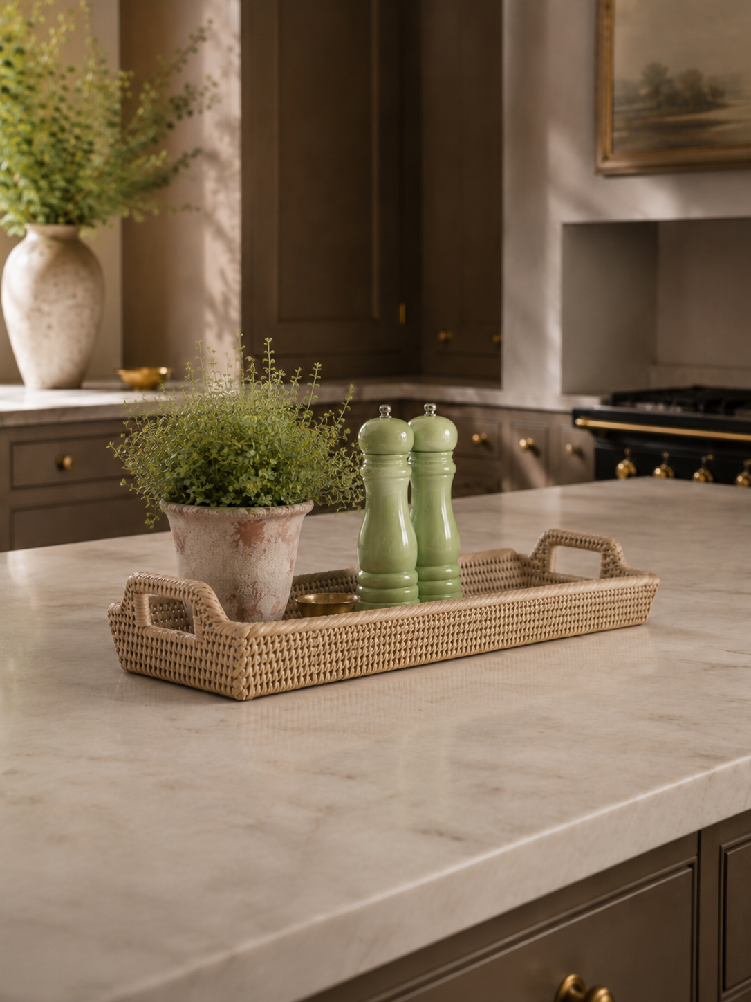 A rectangular woven rattan tray with loop handles sits on a light marble kitchen island, styled with a small potted green plant and two light green pepper and salt mills. The background shows dark brown cabinetry and a stove in a real kitchen environment.