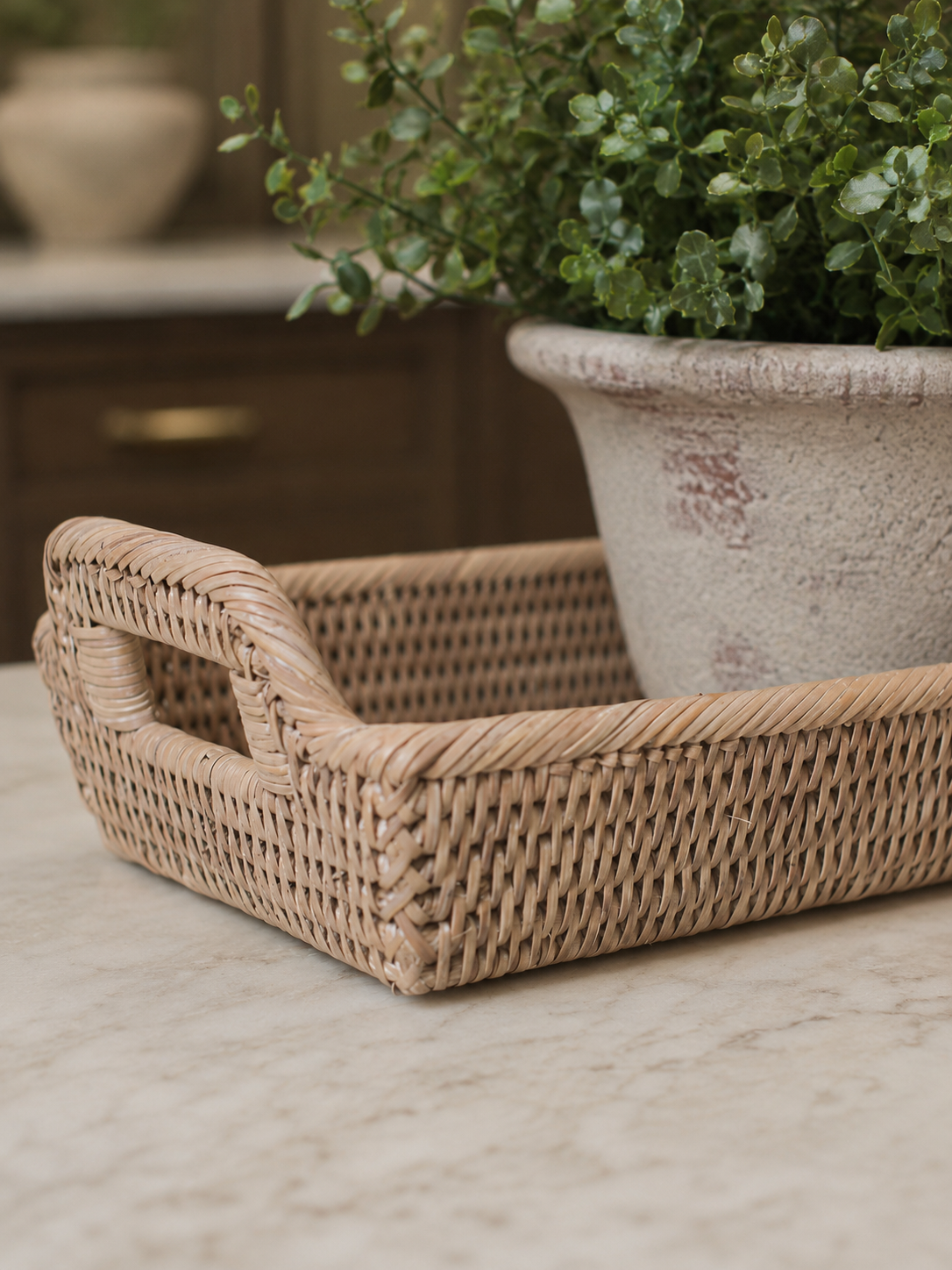 A natural woven wicker storage tray with integrated handles sits on a light marble countertop next to a potted green plant in a rustic ceramic pot.