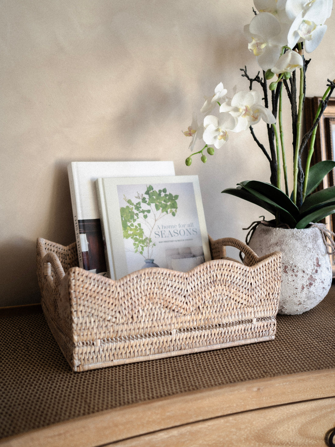 Woven white washed rattan scallop basket tray with books and a vase of flowers on a wooden surface.