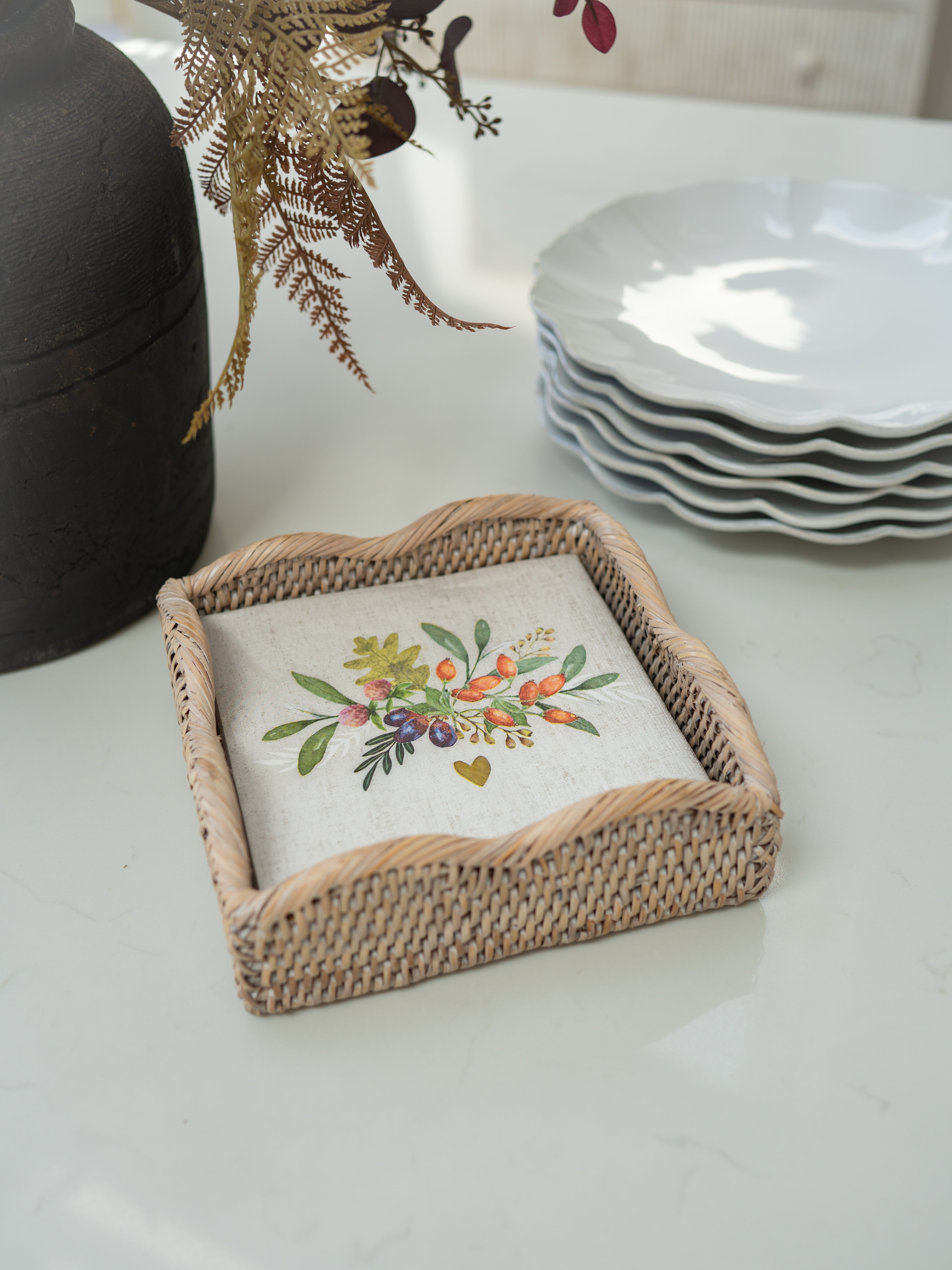 A square woven rattan tray holding a square cream linen napkin with a colorful floral and berry illustration, styled on a white tabletop with a dark vase, dried foliage, and a stack of white plates in the background.