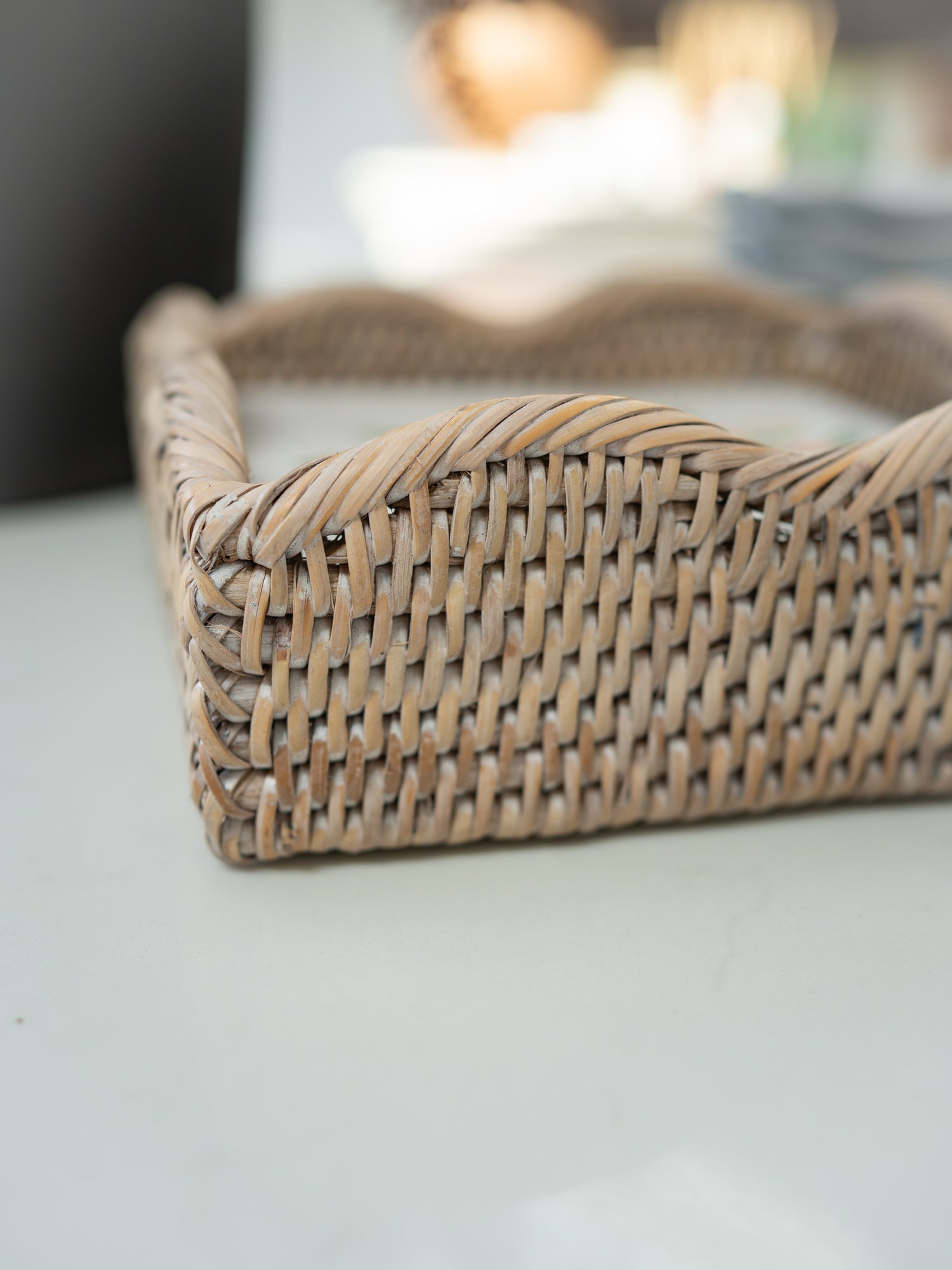 A close-up of a woven natural rattan napkin holder with a wavy scalloped edge, resting on a light-colored surface with a blurred background.
