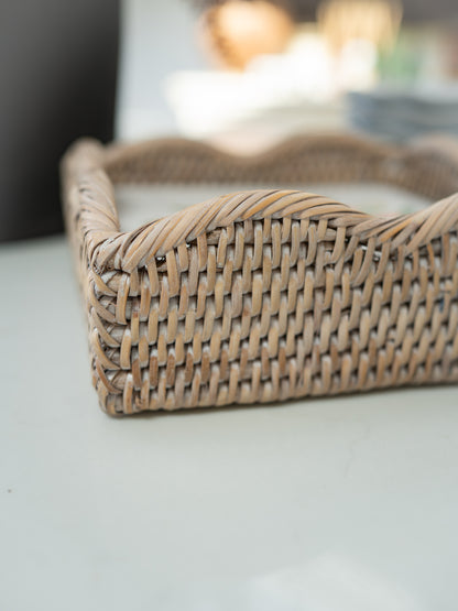A close-up of a woven natural rattan napkin holder with a wavy scalloped edge, resting on a light-colored surface with a blurred background.