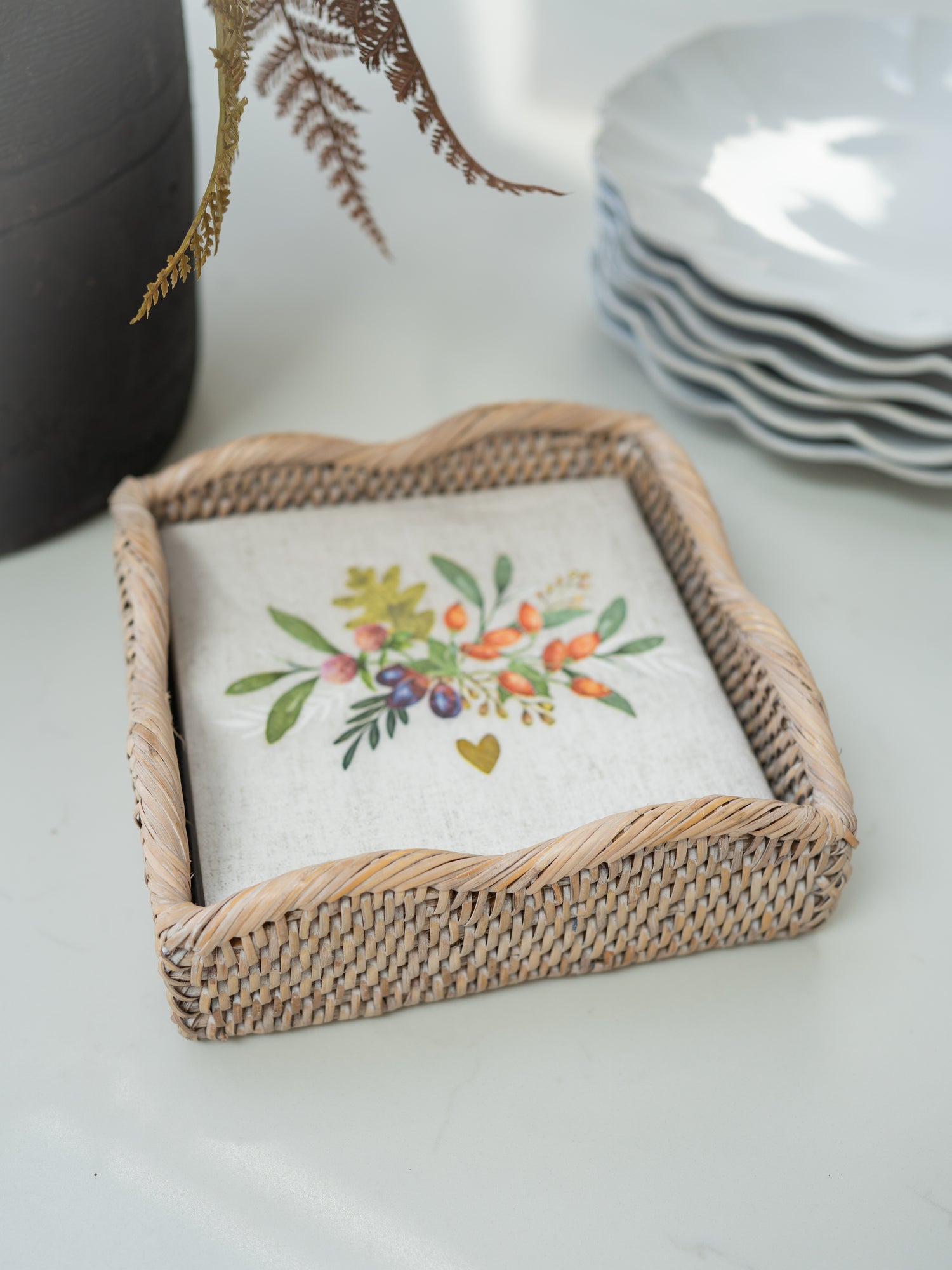 A square woven rattan napkin holder holding a folded cream linen napkin with a colorful floral and berry illustration, set on a white tabletop with blurred plates and a dark vase in the background.