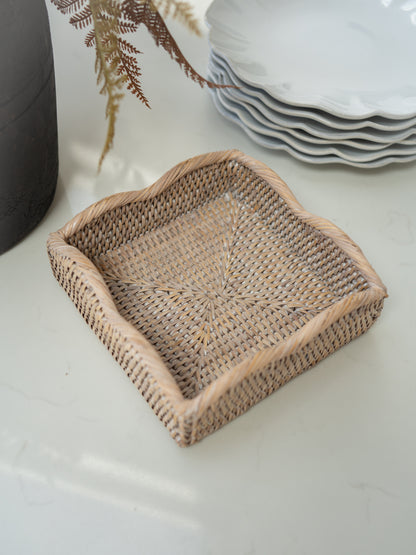A square woven rattan napkin holder with scalloped edges, shown on a light marble tabletop with a stack of white plates and a dark vase in the background.