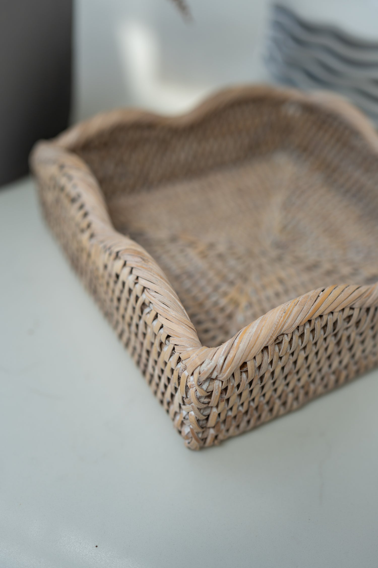 A natural woven rattan napkin holder with a scalloped edge, shown on a light gray surface with a soft-focus background.