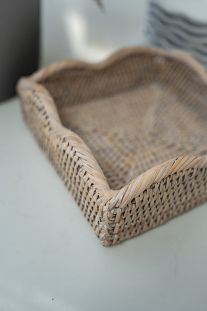 A natural woven rattan napkin holder with a scalloped edge, shown on a light gray surface with a soft-focus background.