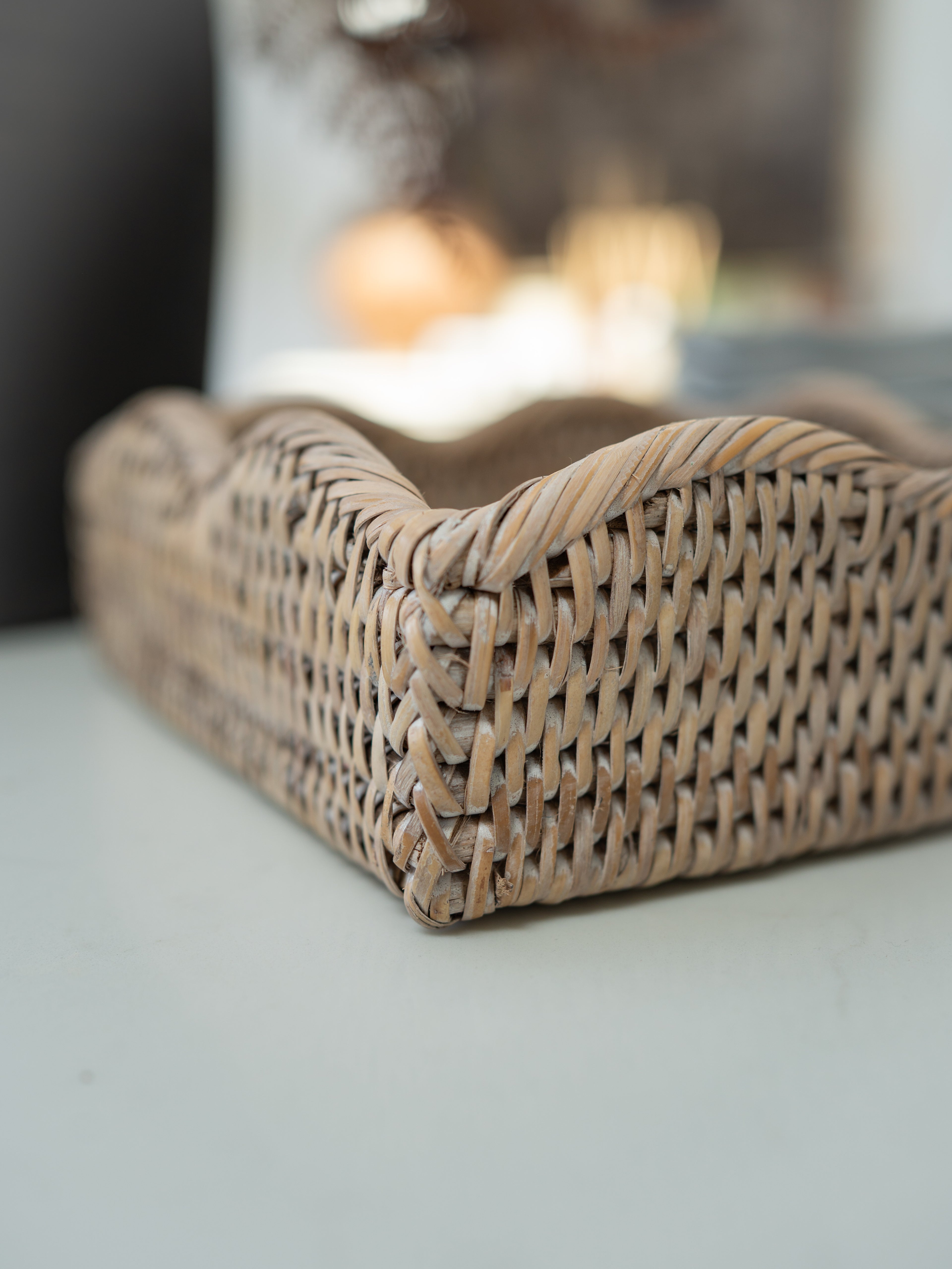 A close-up of a woven natural rattan or wicker napkin holder with a slatted interior, resting on a light surface with a softly blurred background.